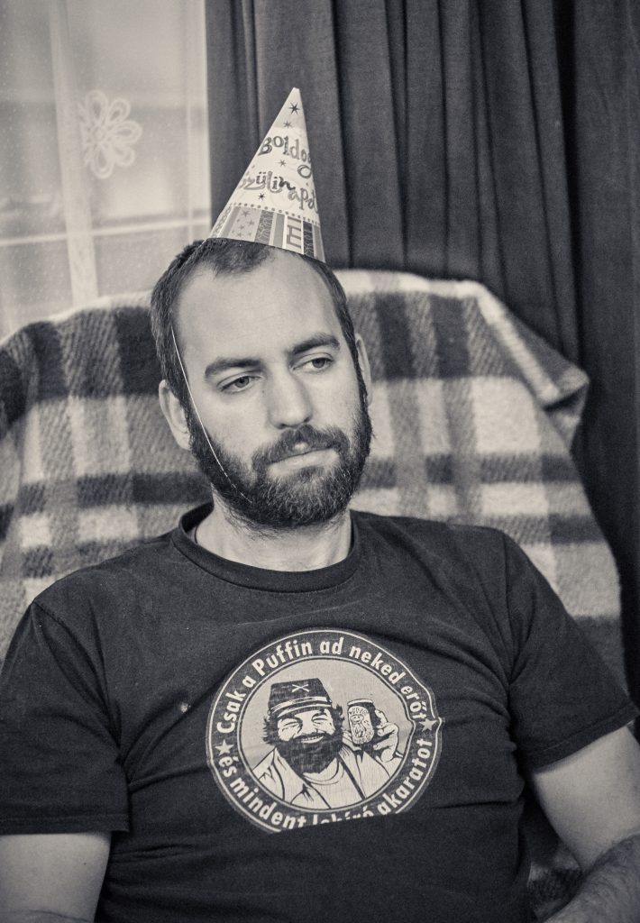 Man in party hat sitting on couch wearing t-shirt with bearded character logo, black and white vintage style photo - © Pascal Recchia. All rights reserved.