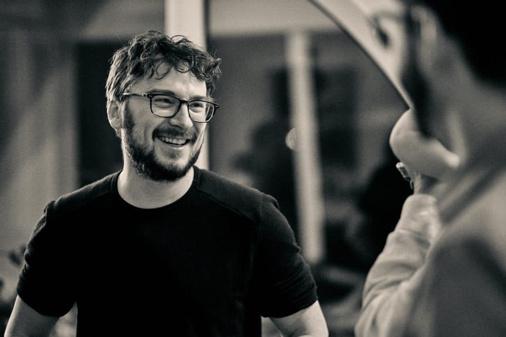 Young man with glasses and curly hair smiling warmly while talking to someone in a casual indoor setting - © Pascal Recchia. All rights reserved.