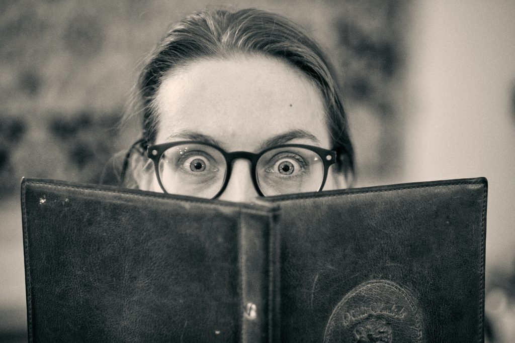 Person with glasses peeking over an open book with wide, surprised eyes in black and white vintage-style photograph - © Pascal Recchia. All rights reserved.