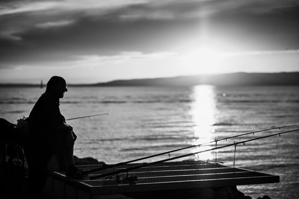 Silhouette of person fishing from dock at sunset with multiple fishing rods, water reflecting golden light - © Pascal Recchia. All rights reserved.