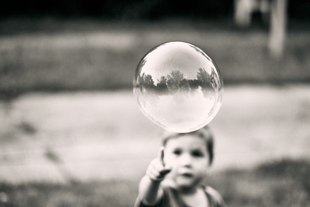 Black and white photo of soap bubble with tree reflections, blurred child reaching toward it in background, vintage film quality