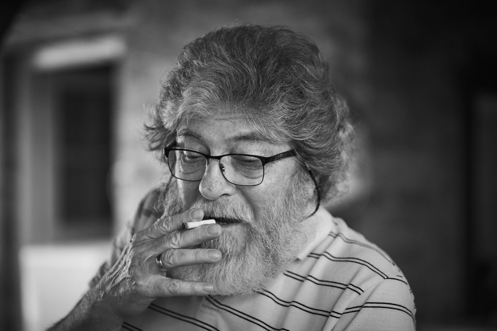Thoughtful older man with glasses and beard smoking, wearing striped shirt in black and white contemplative portrait - © Pascal Recchia. All rights reserved.
