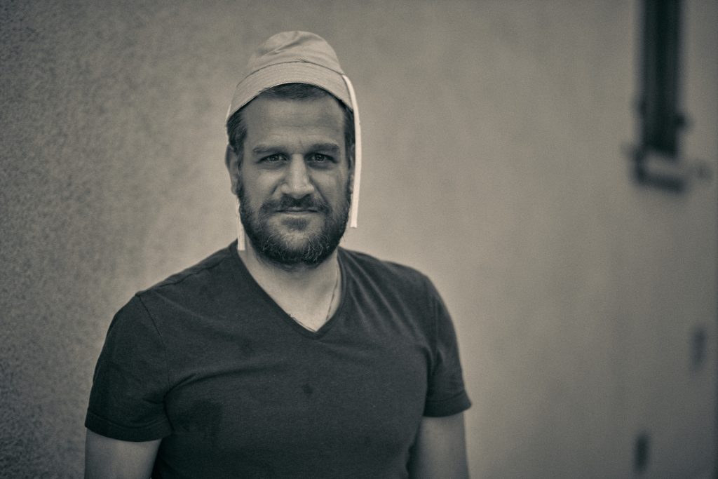 Smiling bearded man in black v-neck shirt and cap against blurred outdoor background - © Pascal Recchia. All rights reserved.