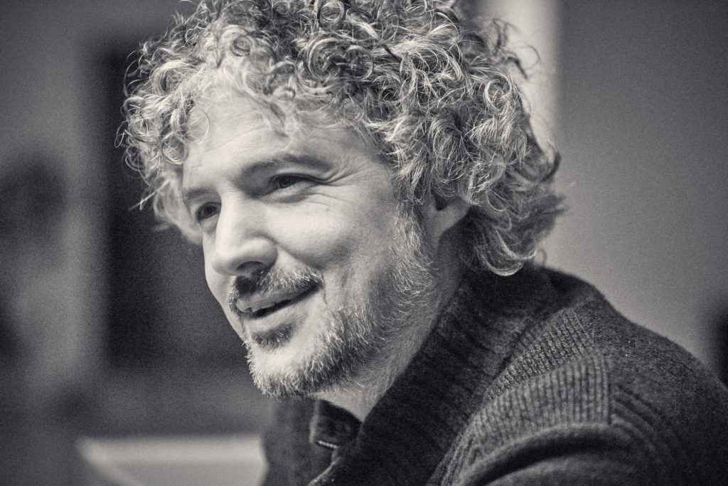 Black and white portrait of smiling man with curly hair and beard wearing knit sweater in warm, intimate lighting - © Pascal Recchia. All rights reserved.