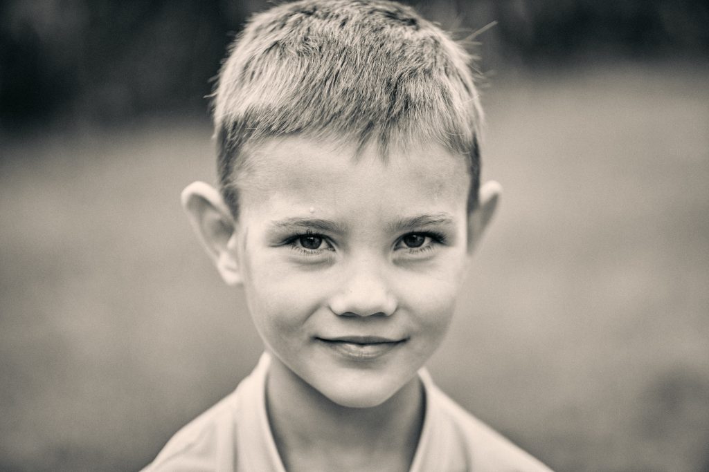 Black and white portrait of a young boy with short hair and bright eyes, smiling softly at the camera in natural light - © Pascal Recchia. All rights reserved.