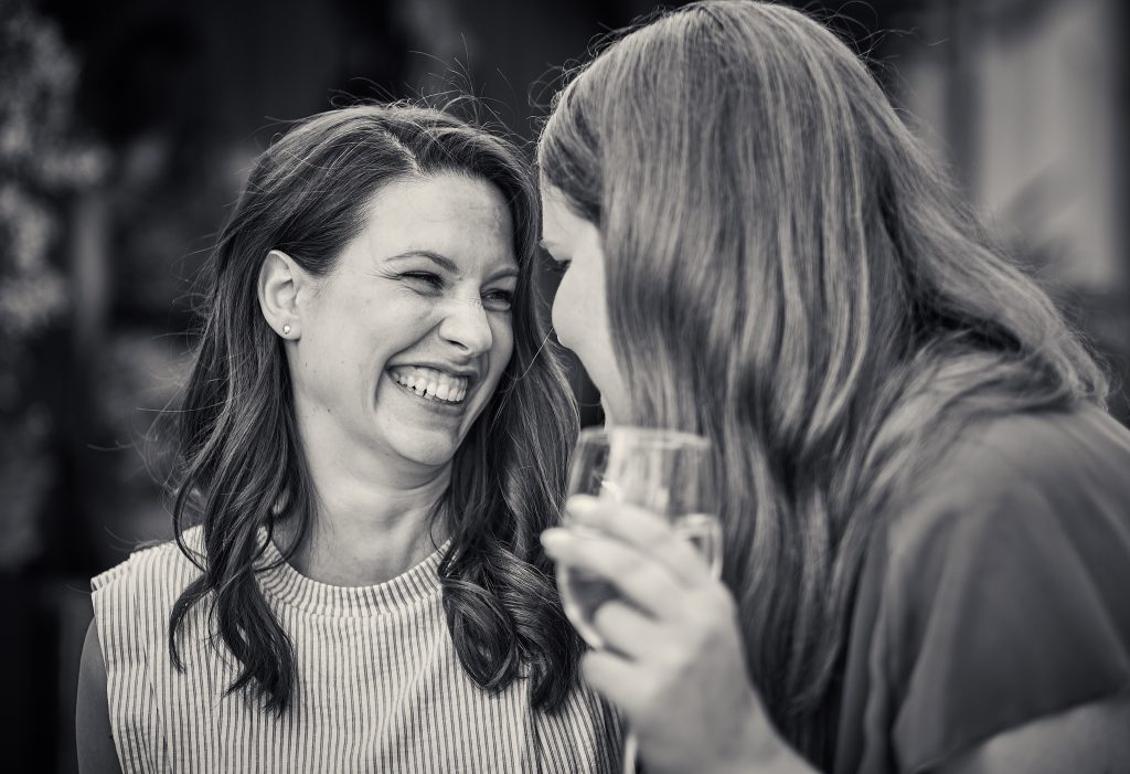 Two women laughing together, one holding champagne glass, black and white photo with warm, joyful moment - © Pascal Recchia. All rights reserved.