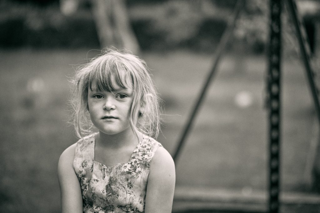 Black and white portrait of young child with tousled blonde hair wearing floral dress, looking directly at camera outdoors - © Pascal Recchia. All rights reserved.
