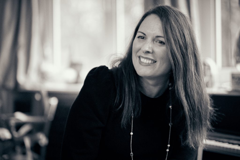 Smiling woman with long brown hair wearing black jacket and necklace in sepia-toned portrait with blurred background - © Pascal Recchia. All rights reserved.