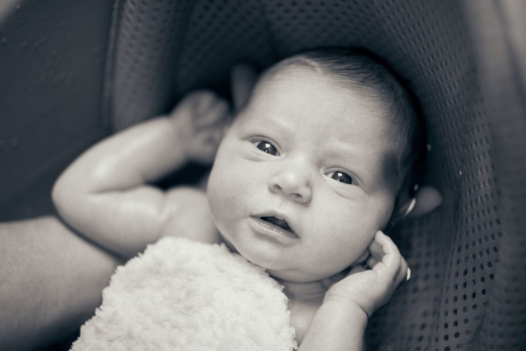 Black and white portrait of baby with dark hair being held, looking at camera - © Pascal Recchia. All rights reserved.