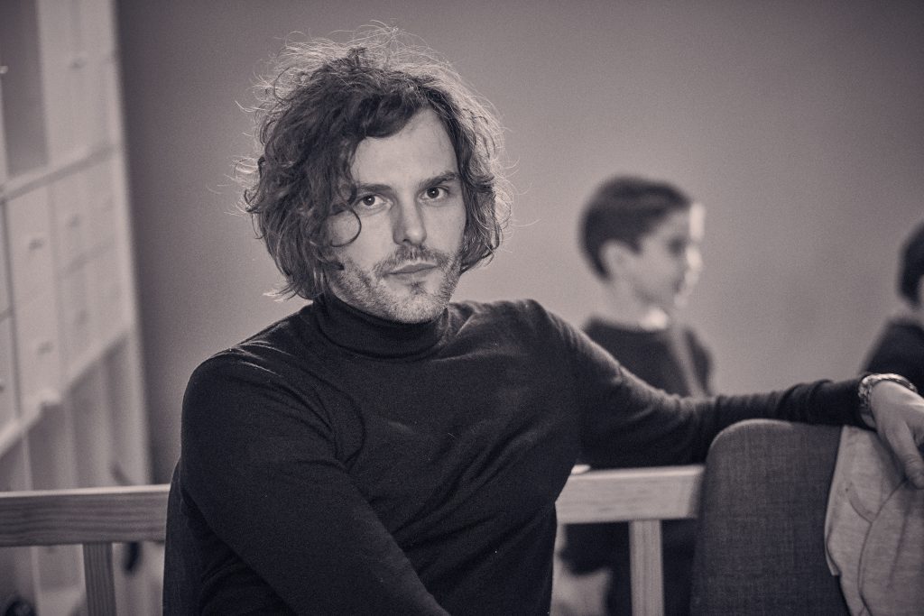 Man with curly dark hair and beard in black sweater sitting at wooden table in warm-toned indoor setting - © Pascal Recchia. All rights reserved.