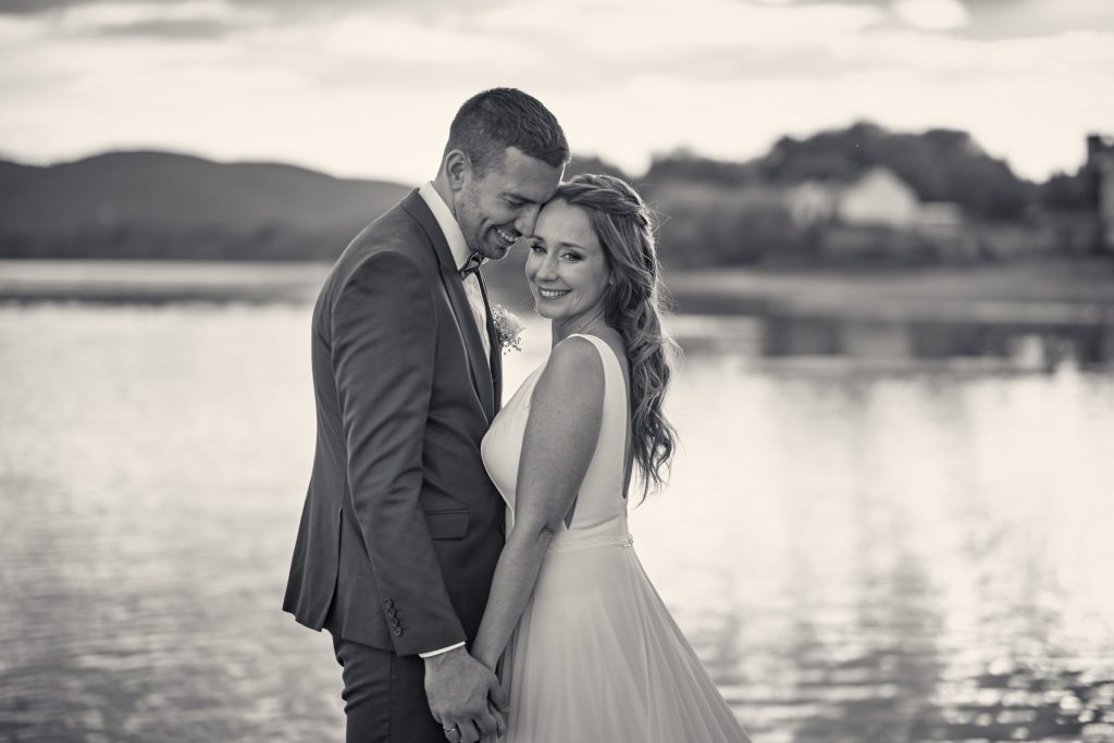 Romantic black and white wedding photo of bride and groom by waterfront at sunset with hills in background - © Pascal Recchia. All rights reserved.