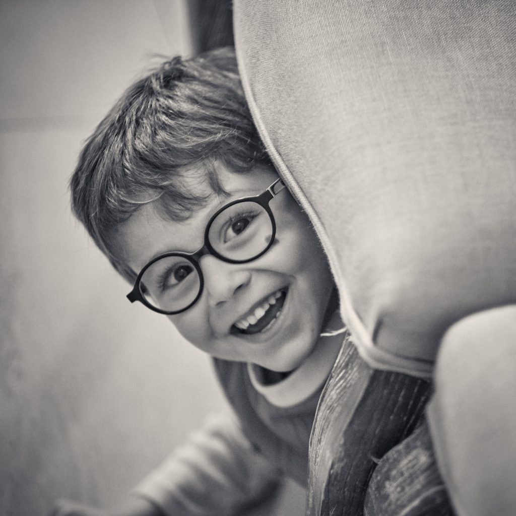 Black and white portrait of smiling young person with round glasses and tousled hair, head tilted playfully to one side