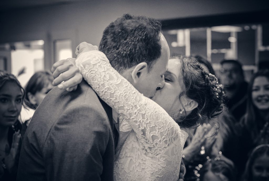 Bride and groom sharing intimate first kiss at wedding reception, black and white photo with guests in background - © Pascal Recchia. All rights reserved.